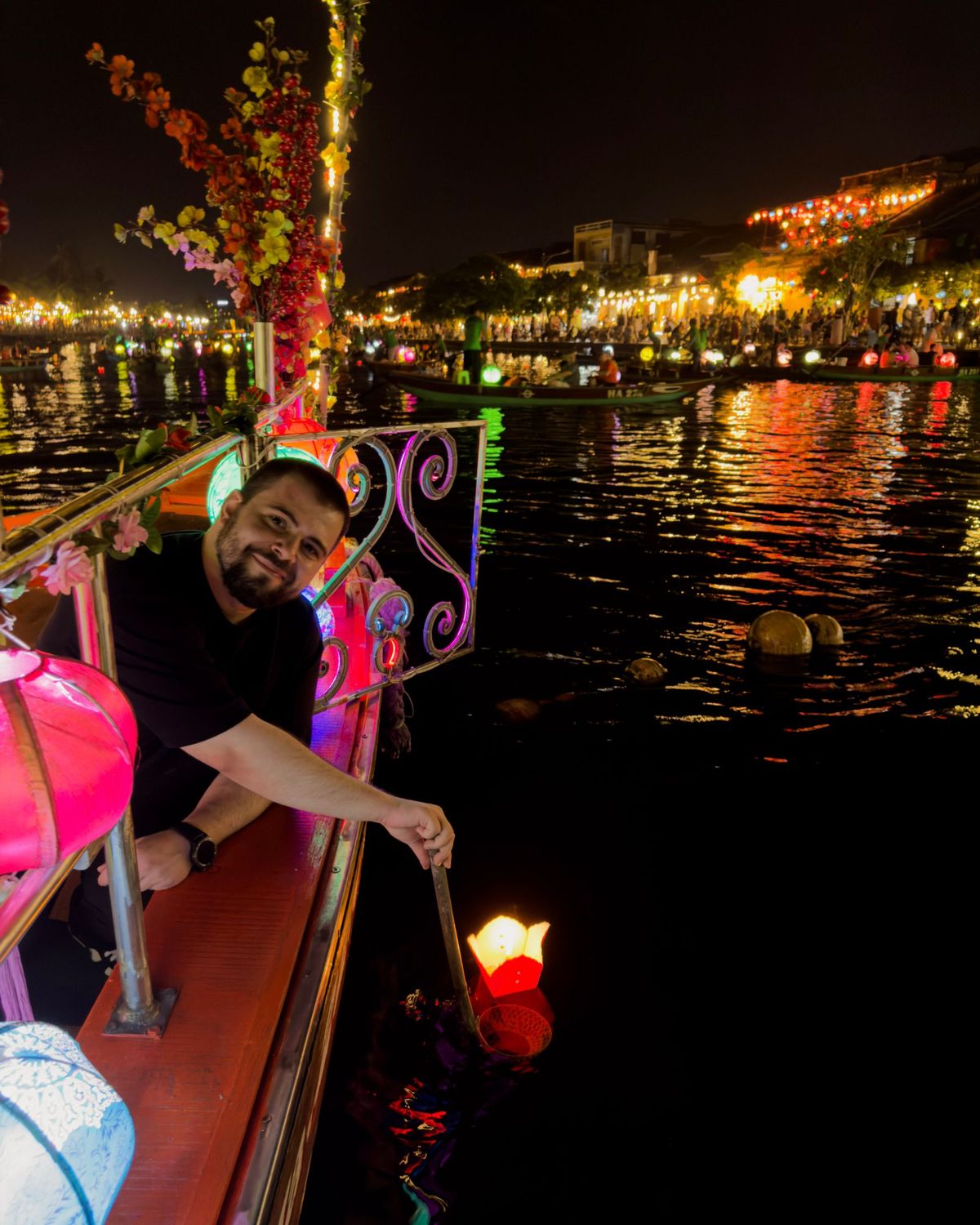 Goksel leaning over the railing of a lantern-lit boat at night, lowering a floating lantern onto the river in Hoi An.