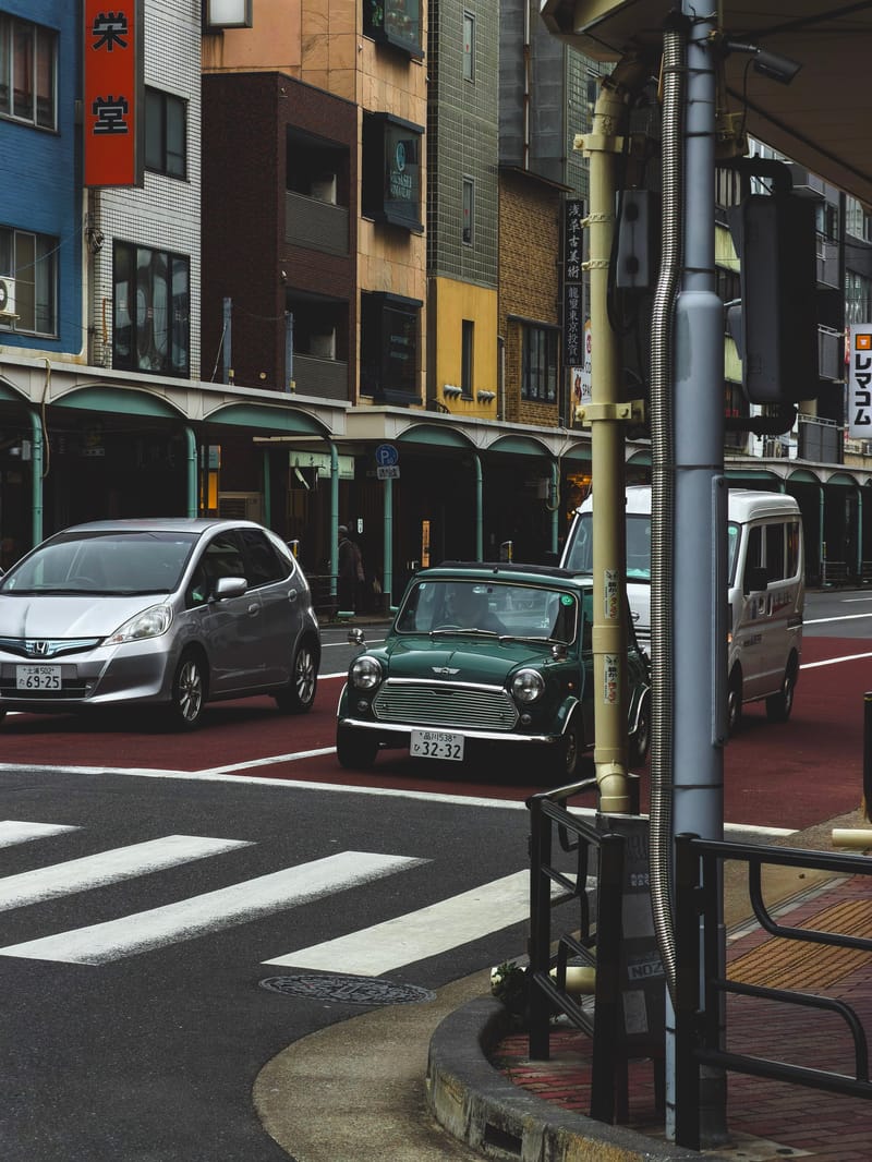 A green classic Mini waits at a crosswalk on a city street, other cars behind it.