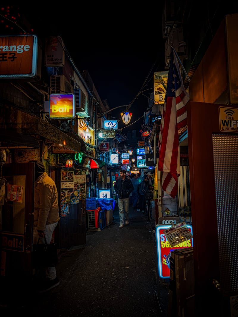 A narrow alley lit by neon signs at night, people walking through.