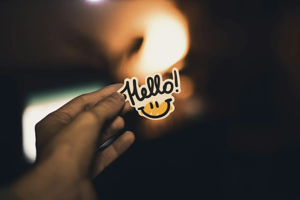 A hand holding a small sticker that says Hello, against a warm blurred light.