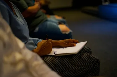 Someone writing in a notebook during a dim talk, with other people seated nearby.